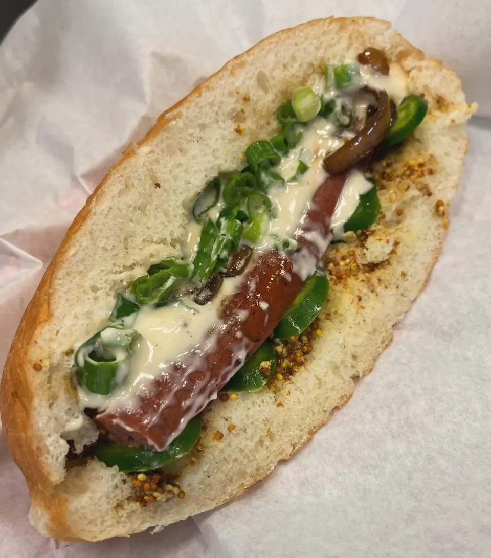 Hot dog in a bun topped with mustard, chopped green onions, relish, and mustard seeds.