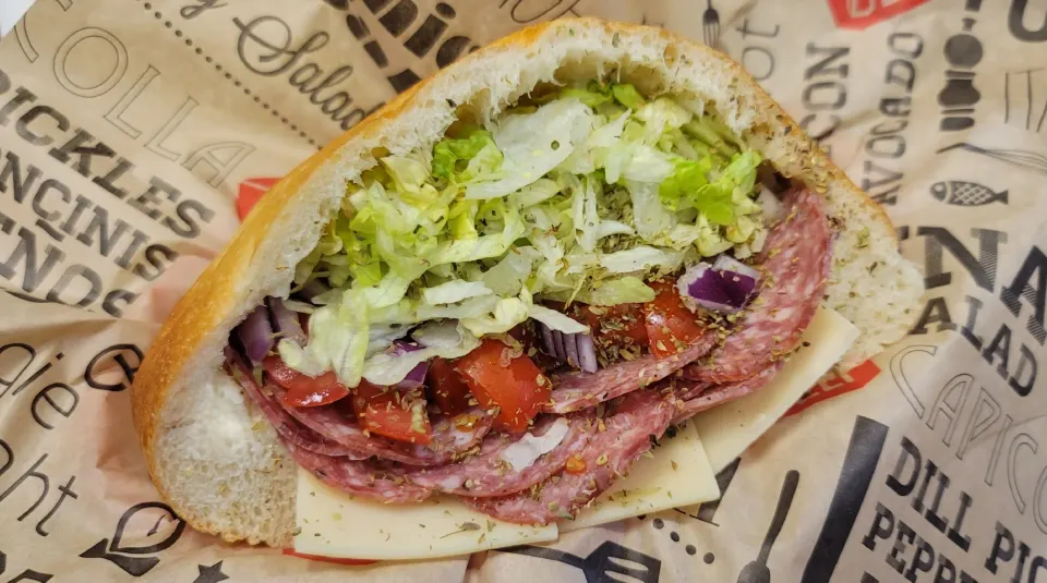 A submarine sandwich with lettuce, tomatoes, onions, slices of cheese, and salami on a piece of paper.
